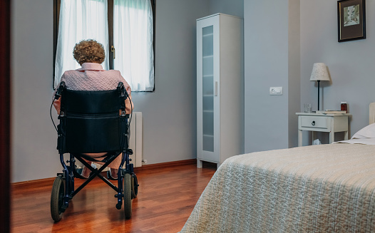 Nursing home resident in a wheel chair left alone, facing a closed window in her room.